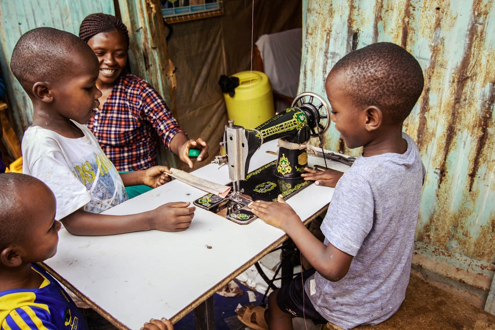 Two children laughing while they learn to sew on a sewing machine with a smiling teacher.